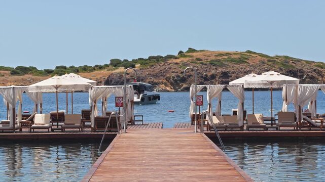 4K view of an upscale beach resort. An empty wooden pier extends into a calm turquoise bay lined with luxury macrame umbrellas and scenic hills.