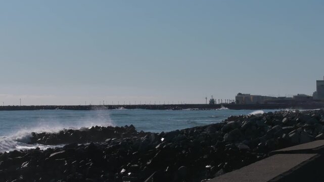 Ocean waves crash against a rocky breakwater near a coastal industrial plant.