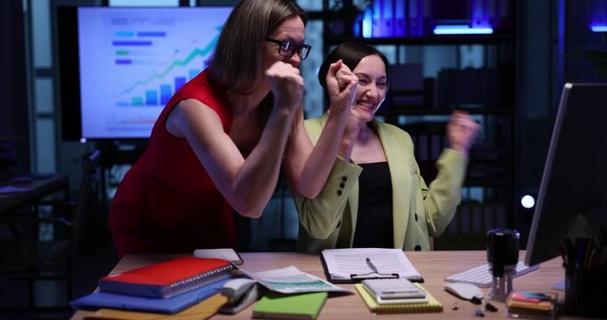 Businesswomen with exited expressions celebrate sales growth in semi-dark office. Traders surprised by extremely rates rising online in workplace