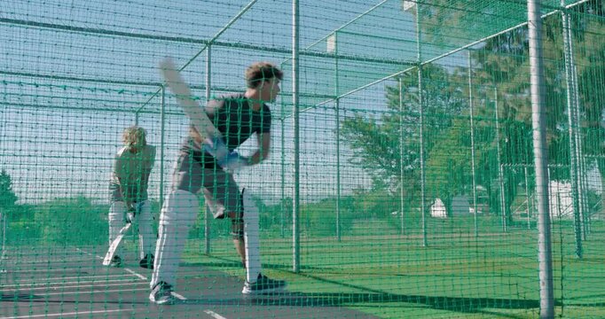 Practicing male cricket partners batting inside green net enclosure on grass pitch, wooden bats