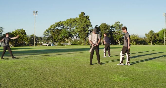 Diverse male friends practicing cricket batting and fielding on outfield, bowler running in