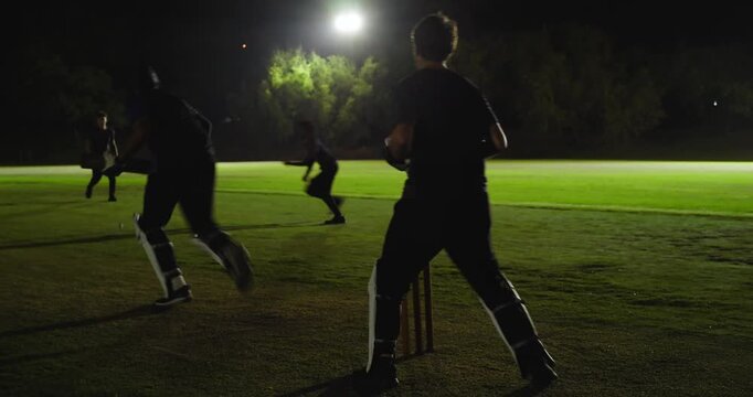 Floodlit cricketers delivering ball past batsman, wicketkeeper diving, fielders retrieving ball
