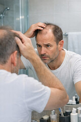 Fototapeta premium Man checks his thinning hair in bathroom mirror. He touches scalp with both hands. He wears white t-shirt. Expression shows concern or worry. Bathroom has glass shower and toiletries