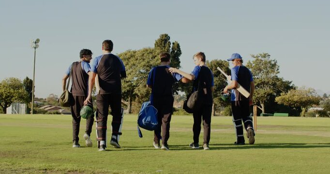 Walking diverse male cricket partners crossing grass outfield, carrying helmets and duffle bags