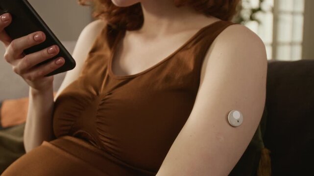 Close up on pregnant Caucasian young adult woman scanning continuous glucose monitor on arm with smartphone. Footage ideal for gestational diabetes awareness and modern prenatal healthcare marketing
