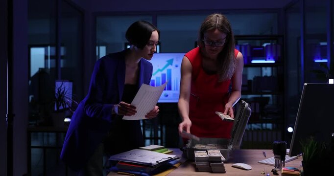 Businesswoman puts money wads into briefcase as assistant holds checklist in dark office. Female entrepreneurs gather cash from deals at desk