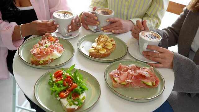 Group of unrecognizable young women chatting and drinking cappuccino in a cafe, enjoying a delicious brunch with a variety of bruschetta toasts served on a modern white table