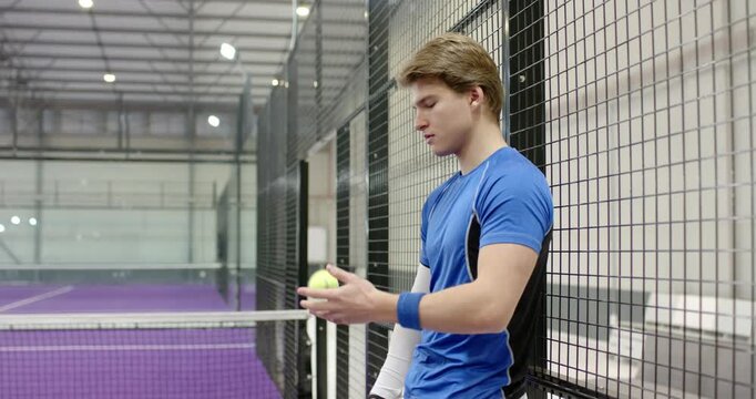 Male athlete leaning on mesh fence, tossing yellow ball over net on padel court and flicking racket