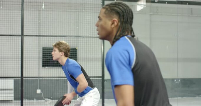 Diverse male teammates tossing ball, swinging paddles on purple court to start rally, copy space