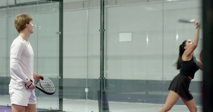 Diverse friends tossing yellow ball, training volley strokes on glass-walled paddle court