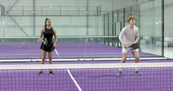 Diverse teammates with rackets serving ball over net on purple padel court, practicing volley