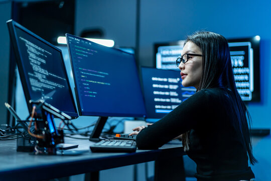 Focused young Asian female software engineer writing code on multiple monitors in a dark modern IT office. Woman programmer analyzing data, cyber security, and artificial intelligence technology