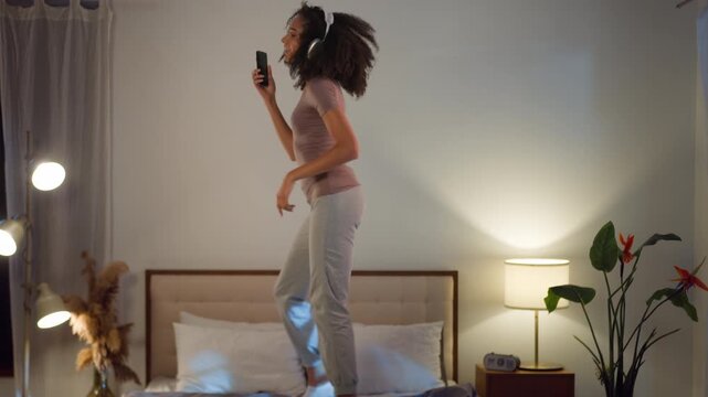 Positive African American woman listening music in headphones jumping dancing on bed at evening. Happy female enjoying singing in bedroom energetic girl jump listen song dance with mobile phone gadget