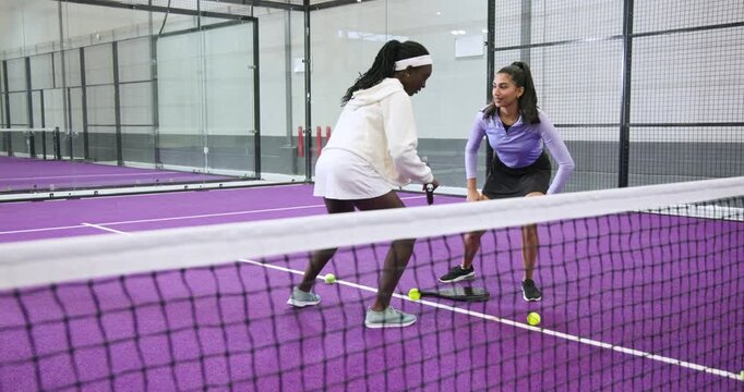 African american coach feeding ball as indian trainee practicing volley drill over net on court
