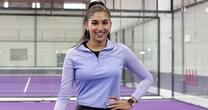 Indian woman entering padel court from right, steadying net, smiling, demonstrating tennis stance