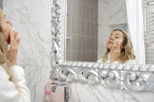 A woman performs her skincare routine in a contemporary bathroom, using cream on her face while looking in an ornate mirror. Soft natural light enhances the clean, aesthetic environment of the space