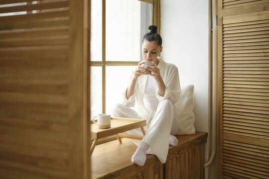 Pretty woman enjoys a peaceful morning, sipping coffee while sitting on windowsill, basking in the natural light