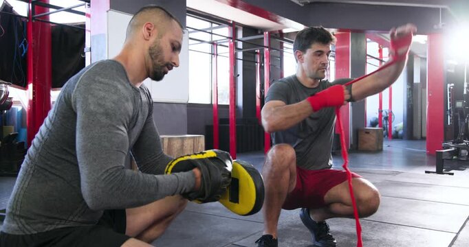 coach raising mitts and trainee boxer wrapping red wrap for pad work at gym
