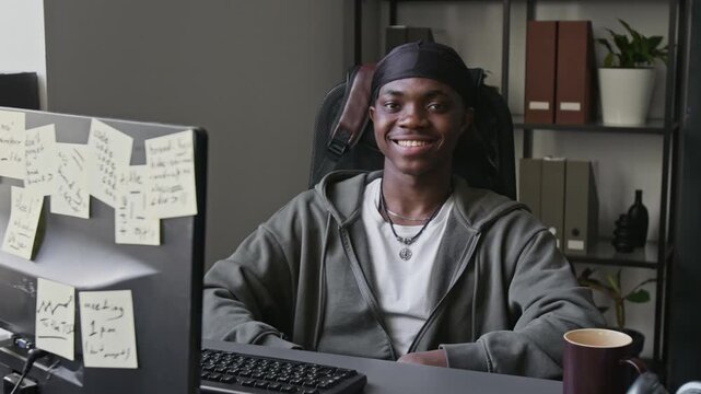 Portrait of African American young man sitting at office desk with computer, turning to look at camera and smiling. Video suitable for tech startups, IT education, or workplace diversity campaigns