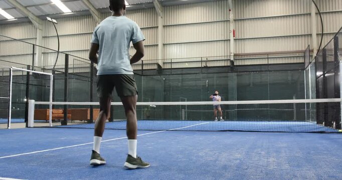 After bouncing padel ball twice for training, diverse male friends serving on padel court