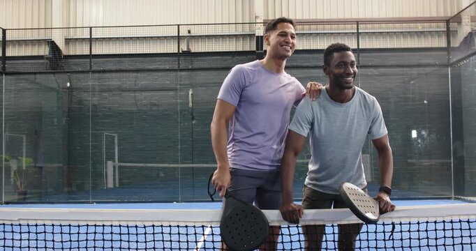 Diverse male friends finishing match leaning on net chatting bumping fists at padel court