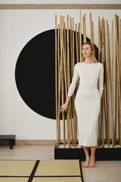 A woman poses next to a bamboo structure in a room with a large black circle on the wall. She wears a fitted dress and stands barefoot on a textured floor