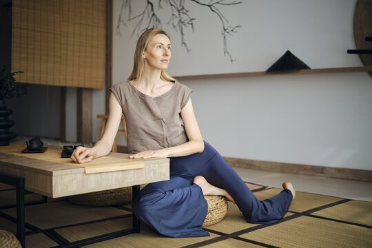 A woman is sitting on a mat in a simple room with a wooden table and minimalist decor. She is focused and calm, holding a cup with a relaxed posture. Natural light fills the space