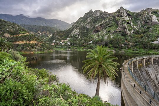 Landscape on La Gomera. The Embalse de Encantadora reservoir. The dam wall on the right. Mountains and some houses in the background. Cloudy sky. La Gomera, Canary Islands, Spain