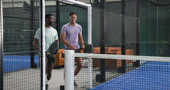 Male padel players entering court with paddles by sofas, fist bumping before volleying ball