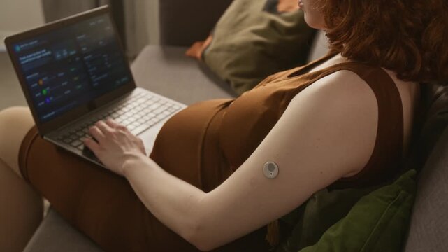 Pregnant Caucasian young adult woman with continuous glucose monitor sensor on arm typing on laptop. Clip is perfect for gestational diabetes awareness and digital health tracking promotions