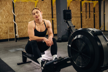 Fototapeta premium Female athlete resting on rowing machine in gym, wearing black sportswear, with wooden wall and fitness equipment visible in the background