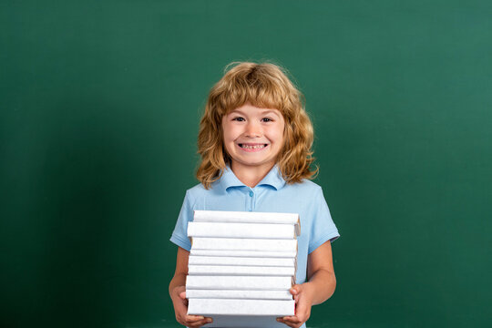 Schoolchild in class. Happy kid against green blackboard. Education and creativity concept. Child holding stack of books with mortar board on blackboard. Back to school