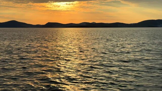A stunning golden sunset over the Aegean Sea in Gulluk, Turkey. Bright sunlight breaks through scattered clouds, casting a warm orange glow over the rippling water and dark mountain silhouettes