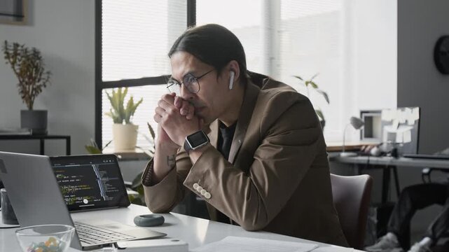 Young Asian man wearing glasses and earbuds typing on laptop, closing it, and working with code on tablet in office. Clip for software development and IT startup commercials