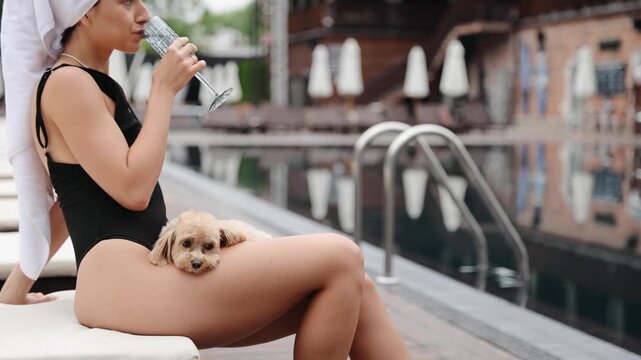 Woman is sitting by the pool at a resort, wearing a swimsuit and having a towel wrapped around her head. She is relaxing with a small dog on her lap and holding a glass of water