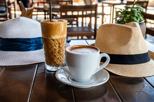 Garden restaurant table with two different coffee orders - hot cappuccino in a mug and cold freddo cappuccino in a glass, along with two straw hats - female and male