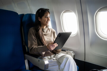 Young woman sitting by the airplane window and using a tablet during a flight, smiling in a comfortable seat. Ideal for travel, business trip, remote work, and modern lifestyle themes. © Thitaporn
