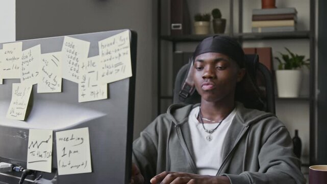Rack focus view of young African American man typing on keyboard while programming at office desk with sticky notes on monitor. Footage for software development, IT education, and tech startup ads