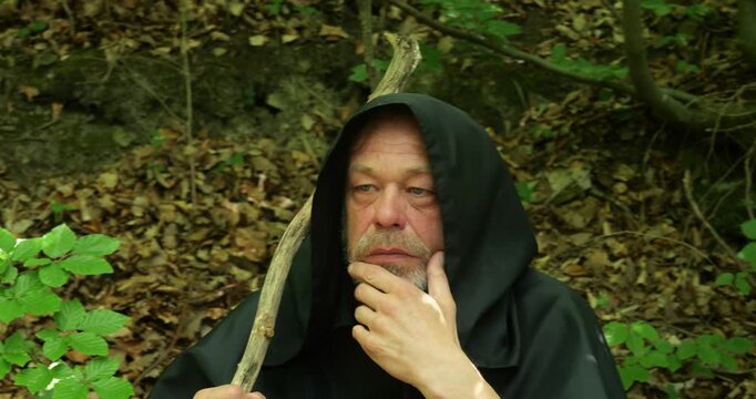 Monk in a dark hooded cloak holds a wooden staff and reflects among dense forest vegetation.