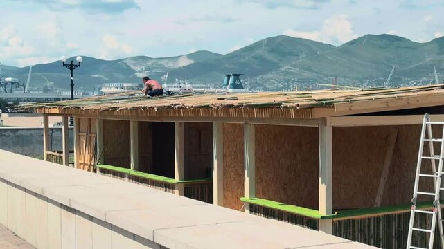 Anonymous handyman laying bamboo poles on wooden kiosk roof near waterfront embankment. Wide daytime shot with streetlamps, commercial ship, and scenic green mountains in the coastal background