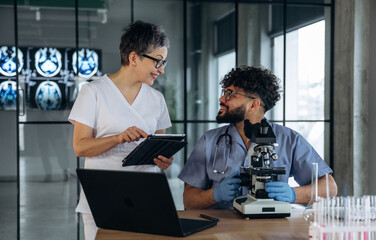 Fototapeta premium Female healthcare professional in white scrubs discusses findings with male colleague in blue scrubs, laboratory setting with microscope and medical imaging in background