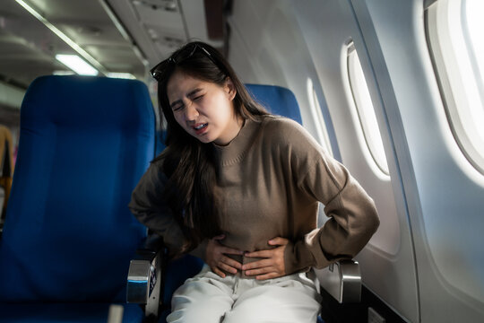Young woman sitting by an airplane window looks unwell and holds her stomach, showing travel sickness or abdominal pain during a flight.