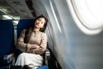Fototapeta premium Young woman sleeping by the airplane window during a flight, sitting in a blue seat with folded arms, calm travel scene with soft light and copy space.