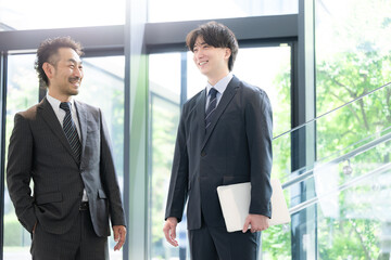 新緑のオフィスの窓際で会話する若手ビジネスマンと上司、取引先のコミュニケーション商談や指導ojtなどの仕事イメージの全身 © kapinon