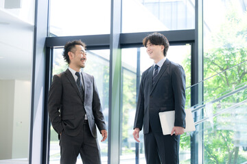 新緑のオフィスの窓際で会話する若手ビジネスマンと上司、取引先のコミュニケーション商談や指導ojtなどの仕事イメージの全身 © kapinon