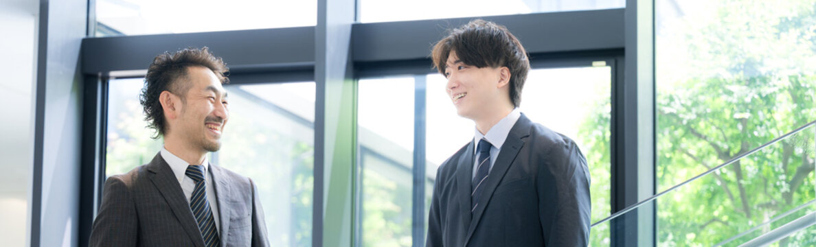 新緑のオフィスの窓際で会話する若手ビジネスマンと上司、取引先のコミュニケーション商談や指導ojtなどの仕事イメージのバナー