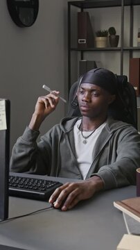 Vertical video of young African American man sitting at desk, twirling pen and looking at computer screen. Footage for IT recruitment and software development marketing campaigns