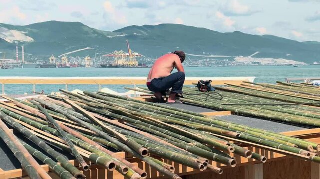 Unrecognizable sunburned shirtless roofer installing bamboo poles on pavilion top with pneumatic nailer. Seaside shelter construction with industrial port and green mountains behind. Medium shot.