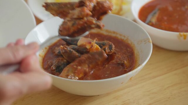 A person scooping and serving sardines in tomato sauce at a dining table