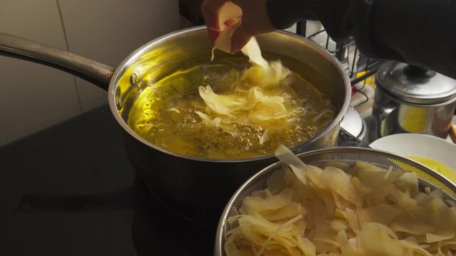 person scooping thinly sliced raw potatoes by hand and placing them into a pot of hot oil for deep frying